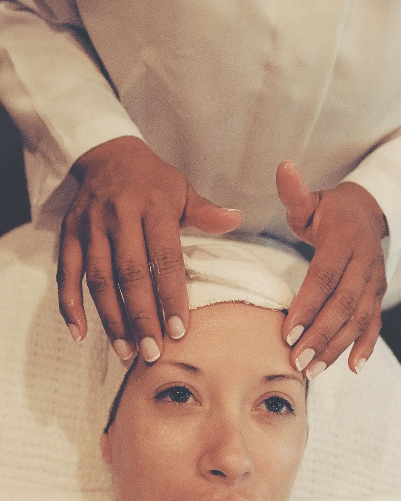 Esthetician's hands resting on the face of a client wrapped in a white towel, ready for a skin treatment