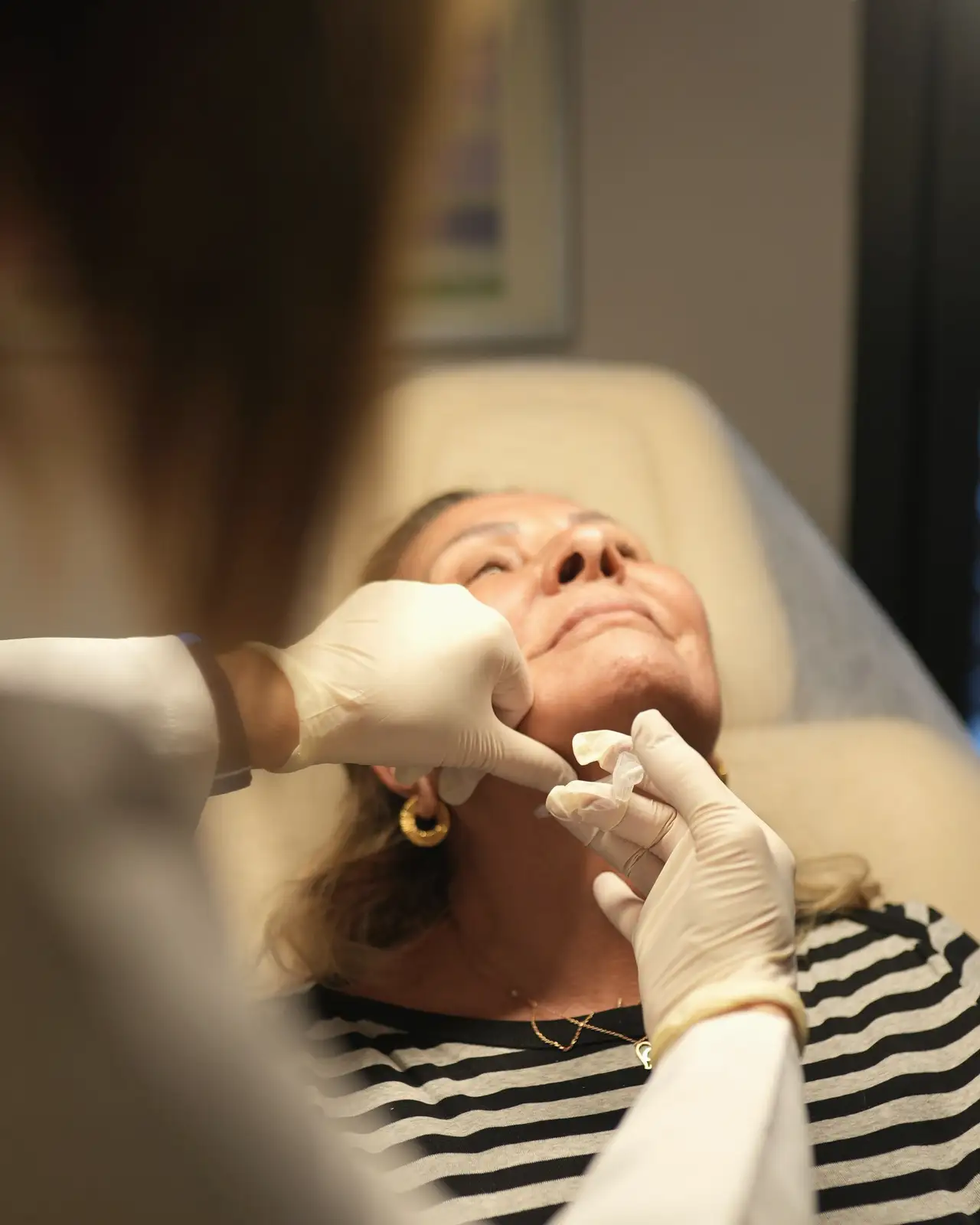 Gloved hands of a nurse injector treating the forehead of a woman lying back on a treatment chair